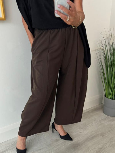 Person in a black top and brown wide-leg pants takes a mirror selfie. They wear black pointed heels, standing on a light wood floor near a tall plant.