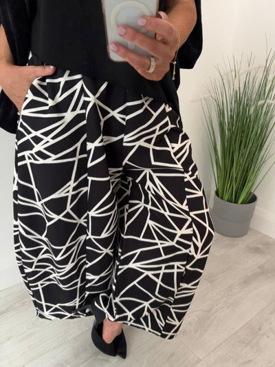 Person wearing black and white patterned pants standing indoors with a plant in the background.
