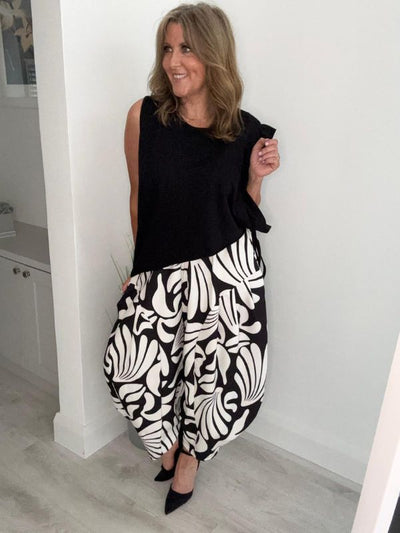 Woman smiling and posing indoors, wearing wide-leg black and white floral patterned pants paired with a sleeveless black top and black pointed-toe heels.