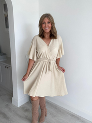 Woman wearing a beige dress with short sleeves and a belt, standing against a white wall.