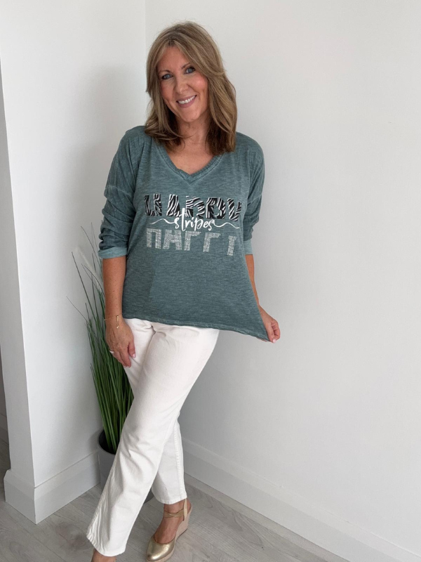 Woman wearing a green graphic t-shirt and white pants standing against a white wall.