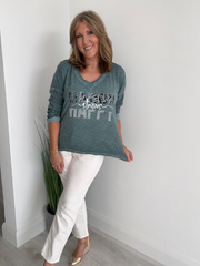Woman wearing a green graphic t-shirt and white pants standing against a white wall.