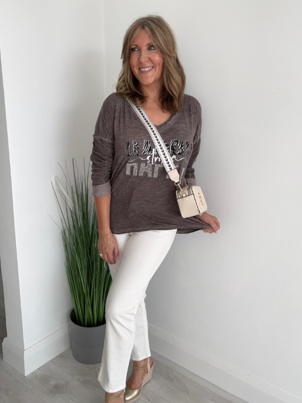 Woman wearing a gray long-sleeve shirt with text and white pants, standing indoors.