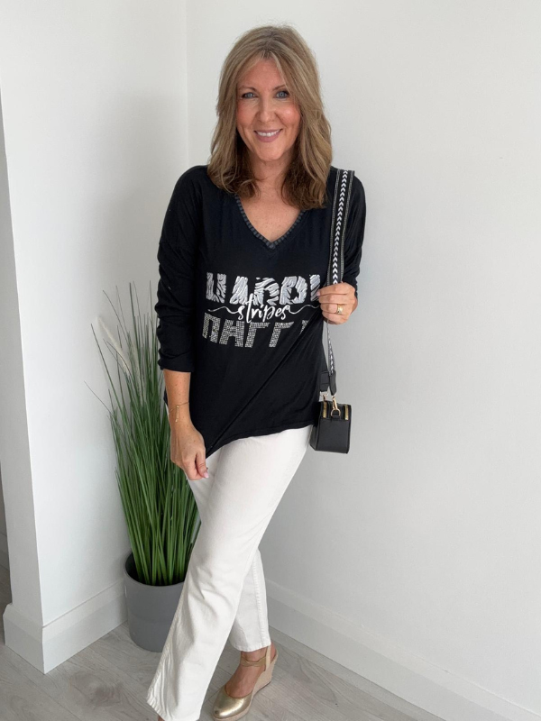 Woman wearing a black sweater with text and white pants, standing indoors.