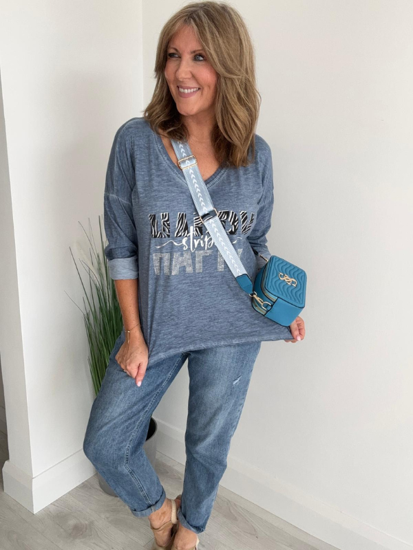 Woman wearing a blue graphic sweatshirt and jeans, holding a blue handbag indoors.