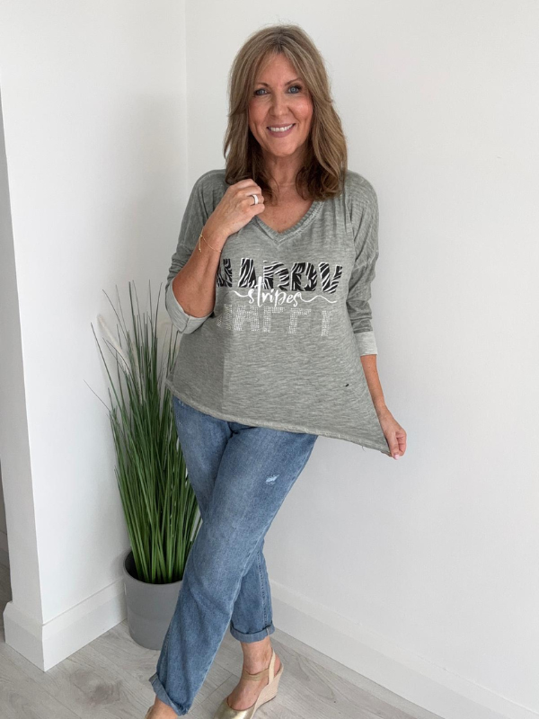 Woman wearing a gray long-sleeve shirt with text and blue jeans, standing indoors against a white wall.