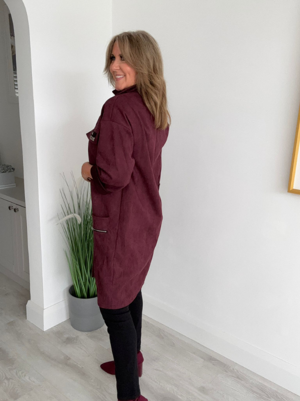 Woman wearing a burgundy coat standing in a room with a white wall and a plant.
