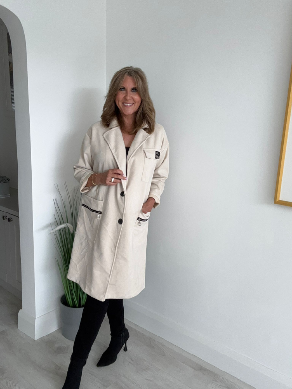 Woman wearing a beige trench coat standing in a room with a white wall and plant.