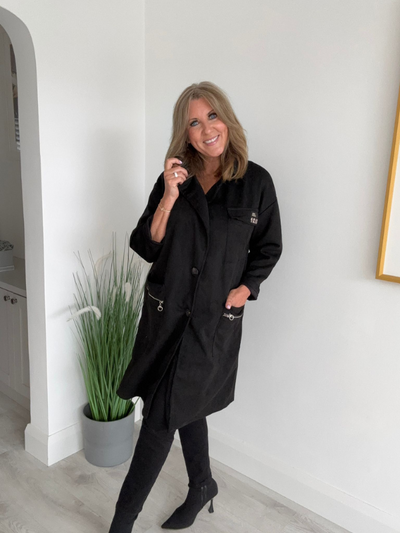 Woman wearing a black coat standing indoors next to a plant.