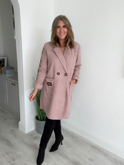 Woman wearing a pink coat indoors with a white wall and plant in the background