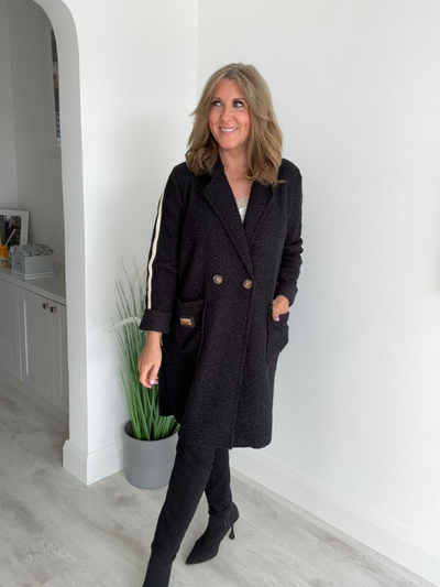Woman wearing a black coat with white stripes standing in a room with a white wall and light-colored floor.