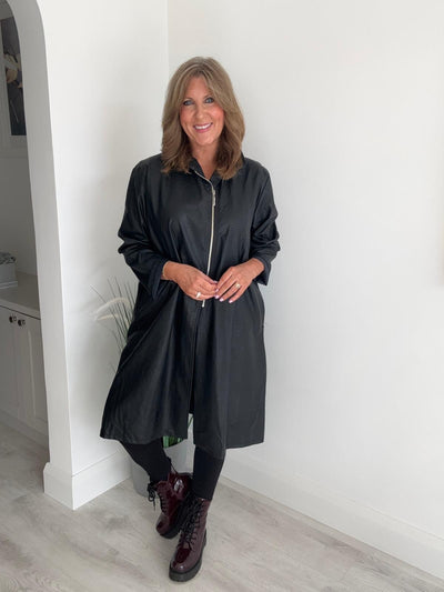 Woman wearing a black raincoat indoors against a white wall.