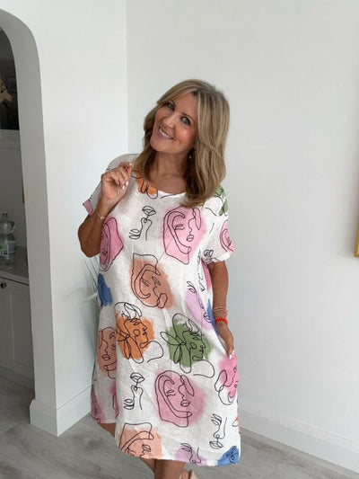 Woman wearing a white linen midi dress with multicolored abstract face print, standing indoors and smiling.