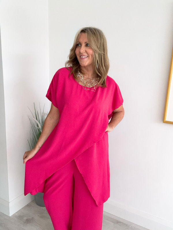 Model wearing an asymmetric co-ord set featuring a short-sleeve top with an uneven hem and matching wide-leg trousers, available in hot pink and blush, designed to fit UK sizes 8–18