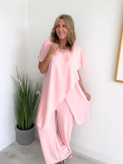Model wearing an asymmetric co-ord set featuring a short-sleeve top with an uneven hem and matching wide-leg trousers, available in hot pink and blush, designed to fit UK sizes 8–18