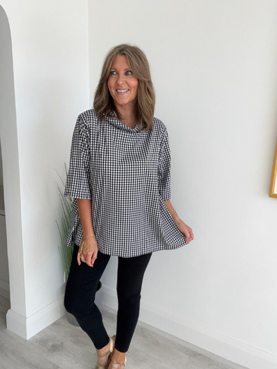 Woman wearing black and white gingham swing top with cowl neck, paired with black leggings and neutral heels.
