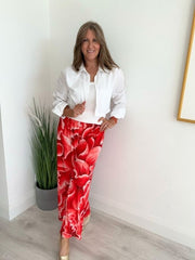 Woman wearing bold red floral wide-leg trousers with white shirt, standing indoors beside a green plant.