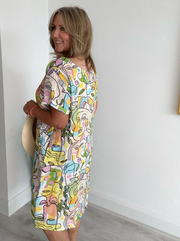 Woman wearing a colorful dress with abstract face designs against a white wall.
