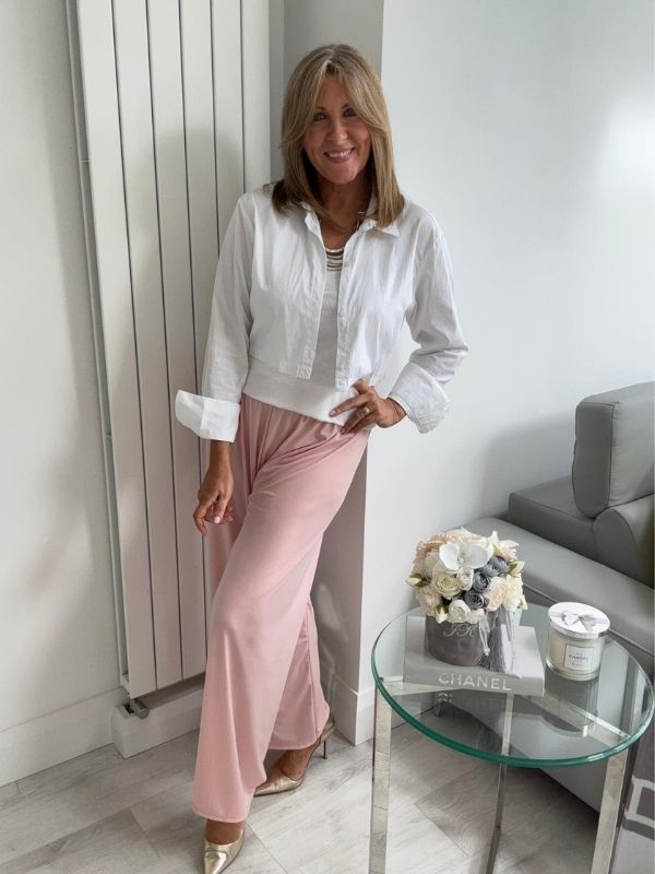 Woman wearing wide-leg slinky pants with a relaxed drape and high-stretch waistband, styled casually with a white shirt and heels, standing in a modern interior space.