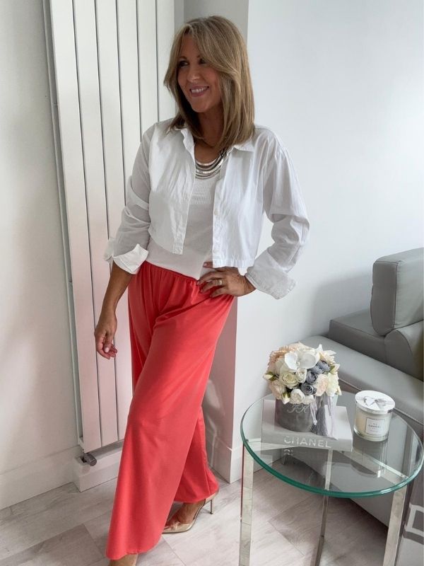 Woman wearing wide-leg slinky pants with a relaxed drape and high-stretch waistband, styled casually with a white shirt and heels, standing in a modern interior space.