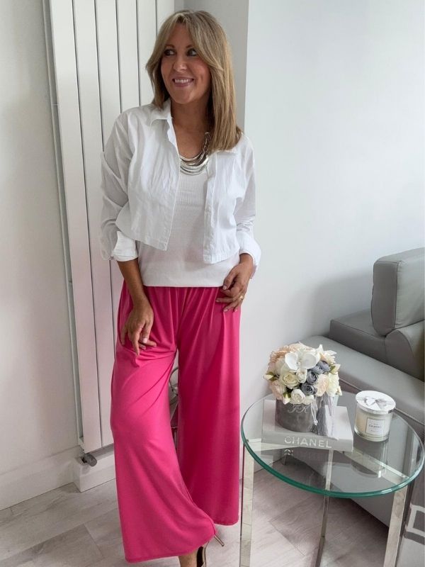 Woman wearing wide-leg slinky pants with a relaxed drape and high-stretch waistband, styled casually with a white shirt and heels, standing in a modern interior space.
