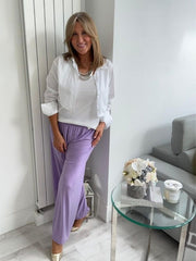 Woman wearing wide-leg slinky pants with a relaxed drape and high-stretch waistband, styled casually with a white shirt and heels, standing in a modern interior space.