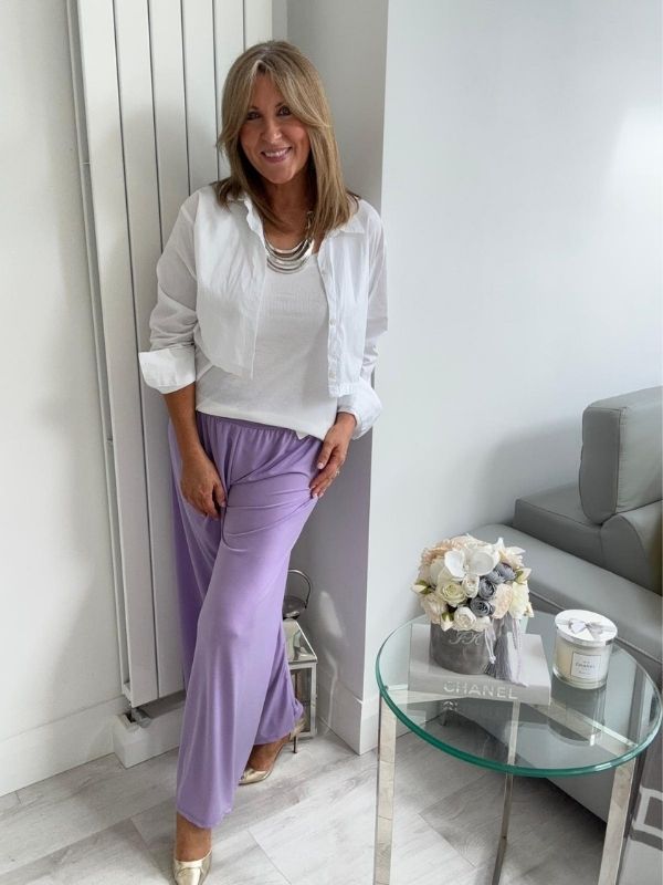 Woman wearing wide-leg slinky pants with a relaxed drape and high-stretch waistband, styled casually with a white shirt and heels, standing in a modern interior space.