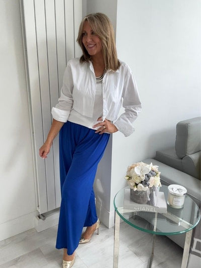 Woman wearing wide-leg slinky pants with a relaxed drape and high-stretch waistband, styled casually with a white shirt and heels, standing in a modern interior space.