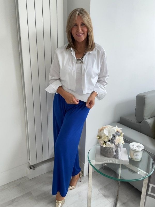 Woman wearing wide-leg slinky pants with a relaxed drape and high-stretch waistband, styled casually with a white shirt and heels, standing in a modern interior space.