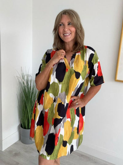 Woman wearing a colorful abstract mini dress with brushstroke patterns in pink, teal, mustard, and white, posing indoors with a cheerful smile.