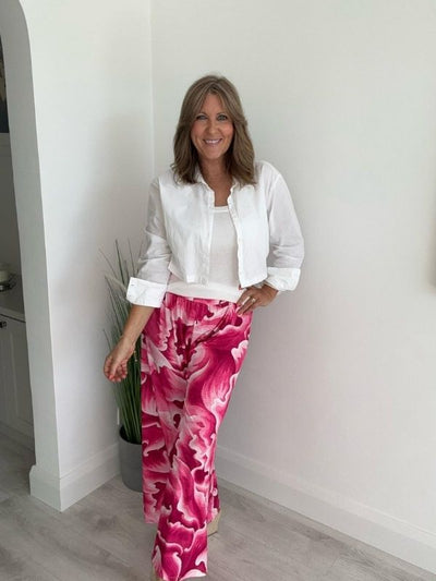 Woman wearing bold Pink floral wide-leg trousers with white shirt, standing indoors beside a green plant.