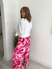 Woman wearing bold Pink floral wide-leg trousers with white shirt, standing indoors beside a green plant.