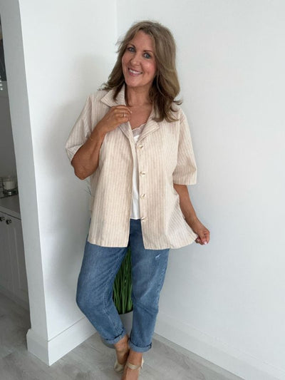 Smiling woman wearing a relaxed-fit beige pinstripe short-sleeve shirt with front buttons, paired with casual blue jeans in a light modern interior.